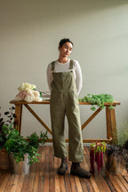 Woman wearing green overalls standing in a room with plants and a wooden table.