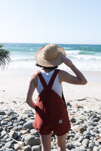 Person wearing a straw hat and red overalls standing on a rocky beach with ocean view