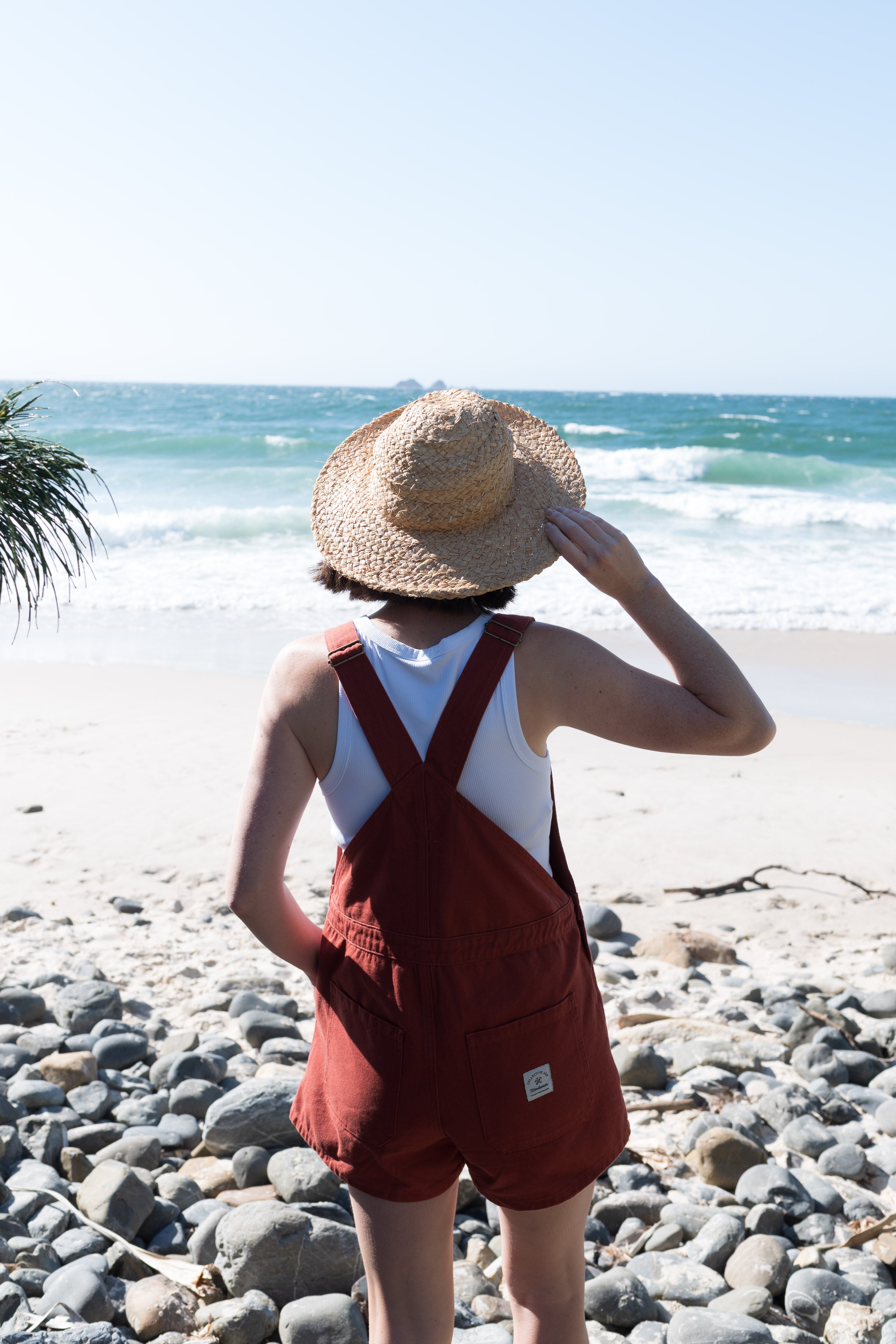 Person wearing a straw hat and red overalls standing on a rocky beach with ocean view