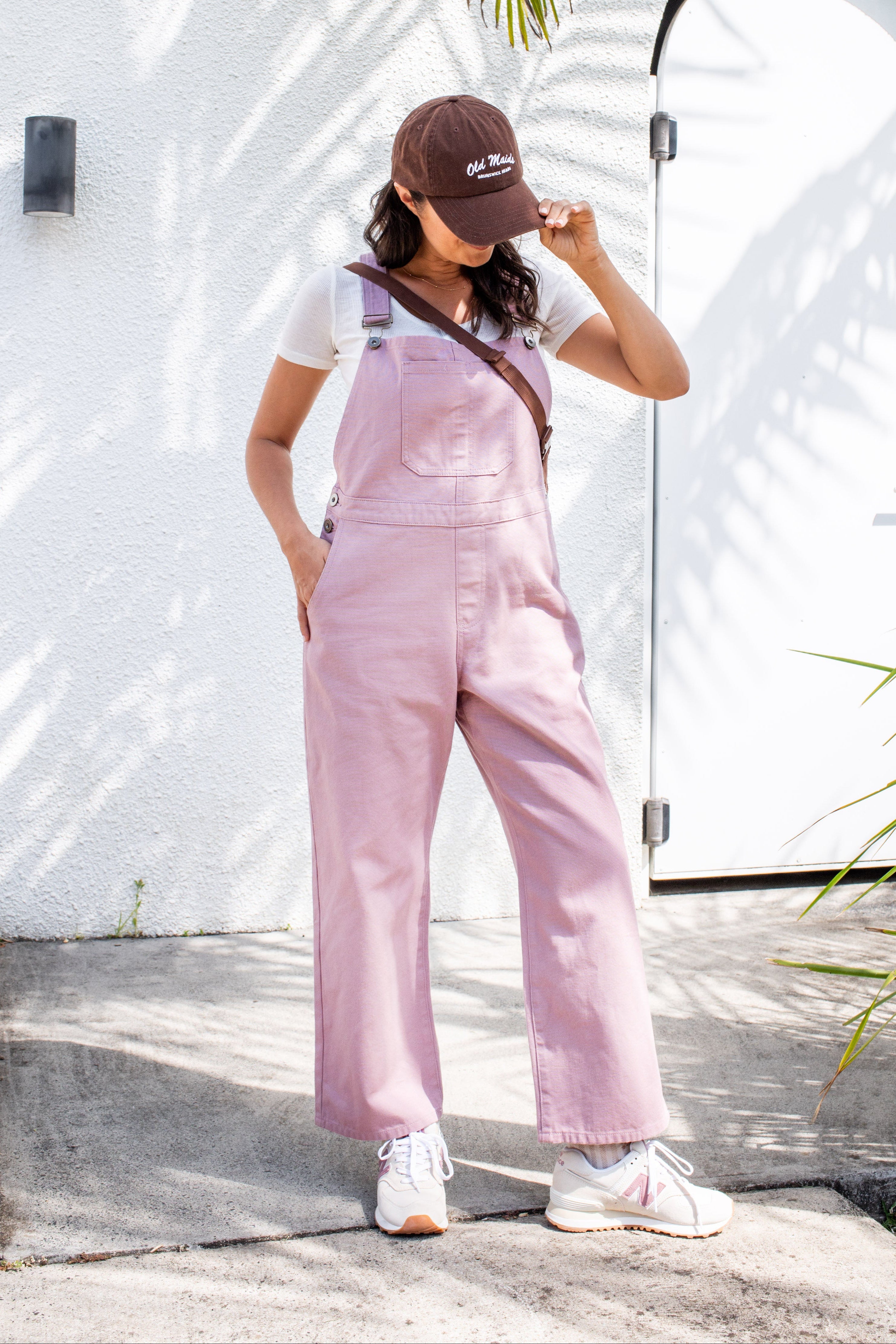 Woman wearing a pink overalls and brown cap standing outdoors with a white wall and palm tree in the background.